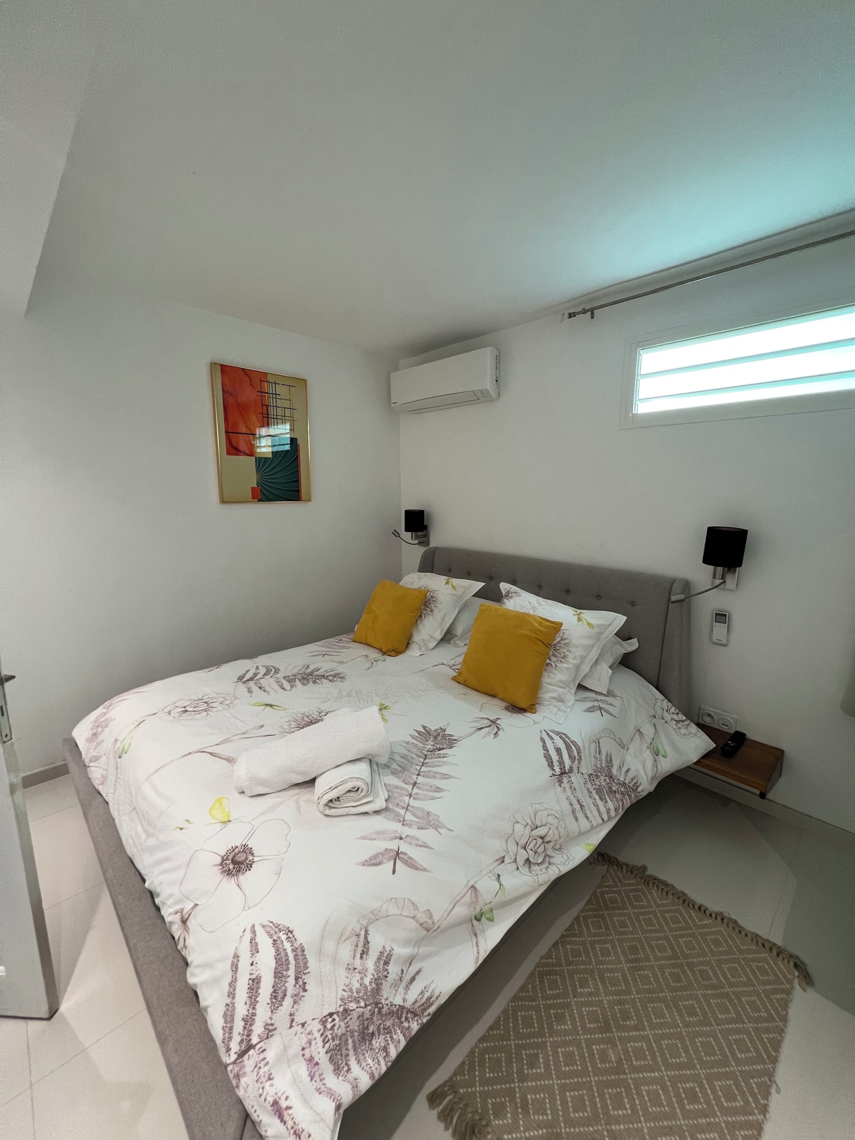 A neatly made bed with floral bedding, a bedside table with a lamp, and a wall-mounted air conditioner.