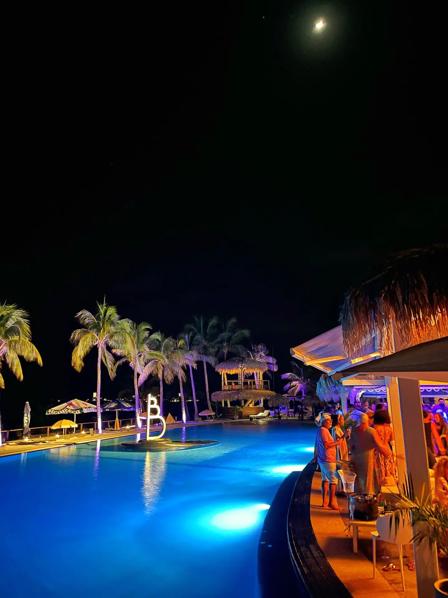 A nighttime pool area with palm trees, a thatched-roof structure, and a 'B' illuminated sign.