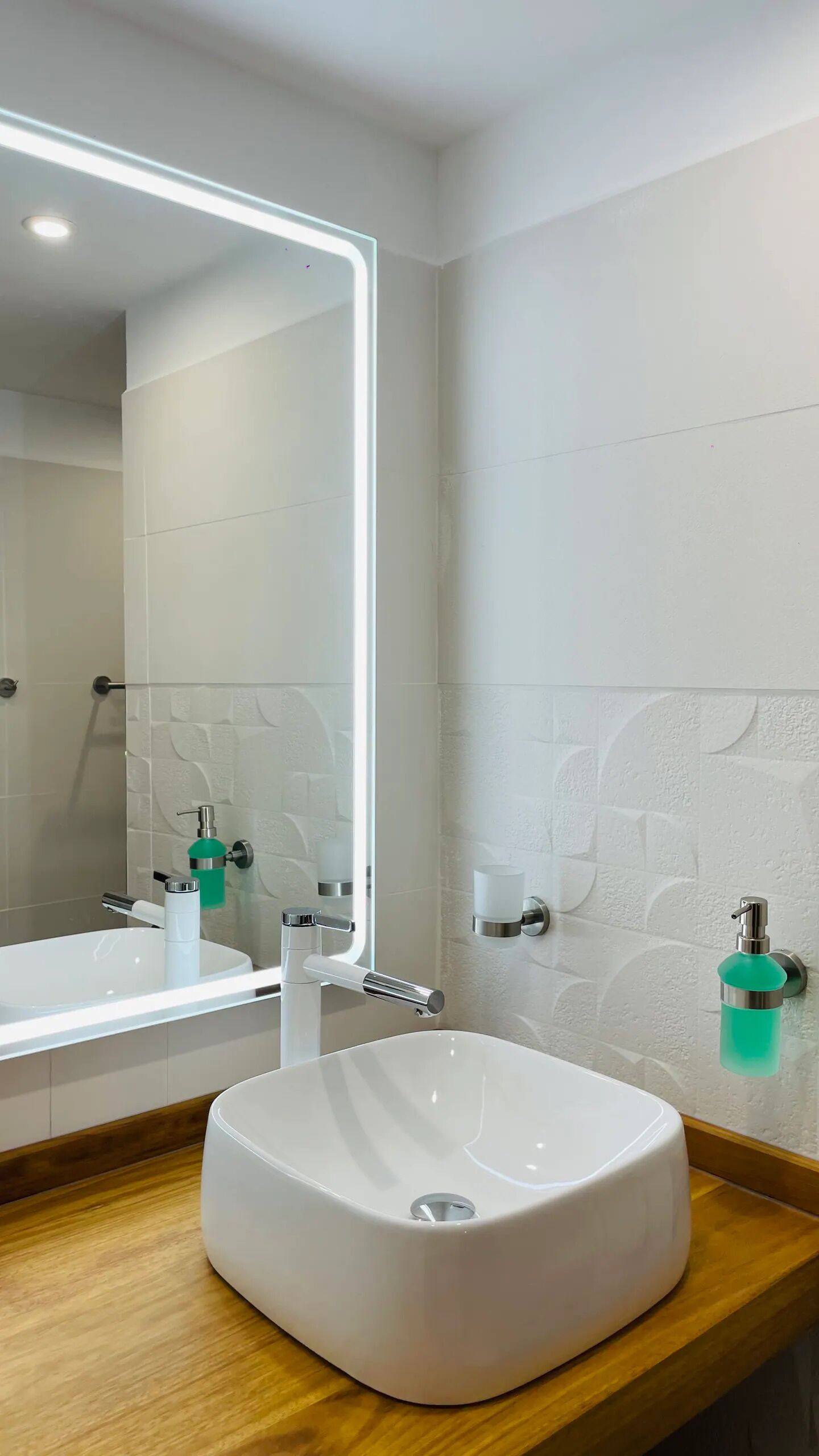 Un baño moderno con un espejo grande, un lavamanos blanco y un dispensador de jabón verde.