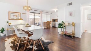 Ein moderner Essbereich mit einem weißen Tisch, Stühlen und einer Kandelaber. Im Hintergrund ist eine Küche mit Holzschränken und einer Arbeitsplatte zu sehen.