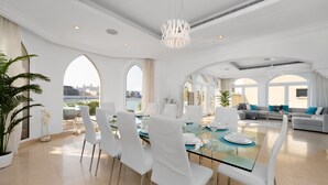 A spacious dining area with a glass table, white chairs, and a chandelier.