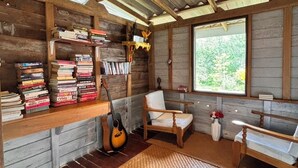 A rustic cabin interior with wooden walls, a bookshelf, a guitar, a chair, and a large window. - Baansuan Suksangob Farm Stay (Yan Ta Khao)