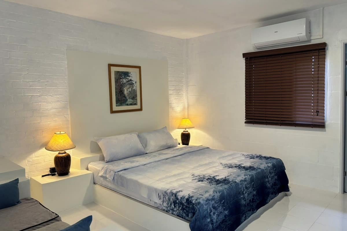 Une chambre à coucher comprenant un lit, deux tables de chevet avec des lampes, un climatiseur fixé au mur et une fenêtre avec des stores.