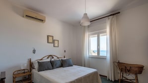 A bedroom with a bed, a window with a view, a wicker chair, and an air conditioning unit. - Seaview Villa In Mongerbino by Wonderful Italy (Santa Flavia)