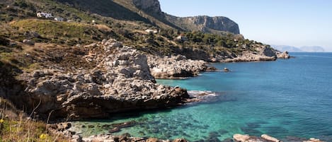 A coastal landscape with rocky terrain, clear blue sea, and distant mountains.