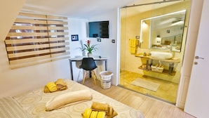 A modern hotel room with a bed, a desk, a chair, a potted plant, and a bathroom with a glass door. - Apartments Ivan (Solin)