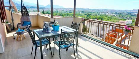 A balcony with a table and chairs, a hanging chair, and a view of a cityscape and mountains.