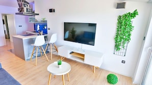 A modern living room with a flat-screen TV, a dining area with white chairs, and a kitchen with a blue cabinet. - Apartments Ivan (Solin)