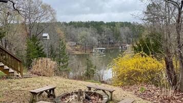 A lakeside picnic area with a fire pit and benches, surrounded by trees and a wooden deck.