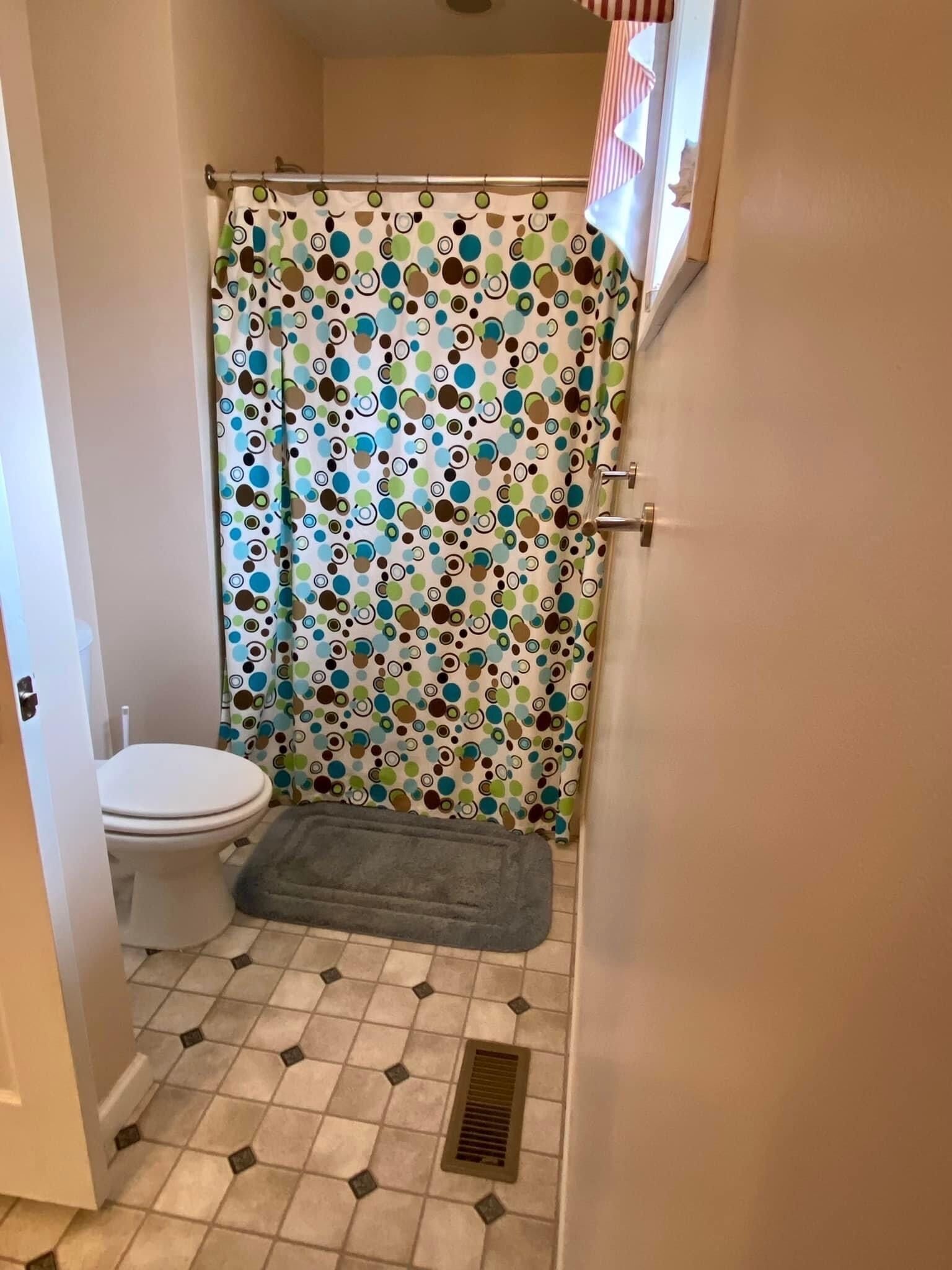 Een kleine badkamer met een toilet, douchegordijn met een kleurrijk rond patroon, een grijze mat en een betegelde vloer met een afvoer.