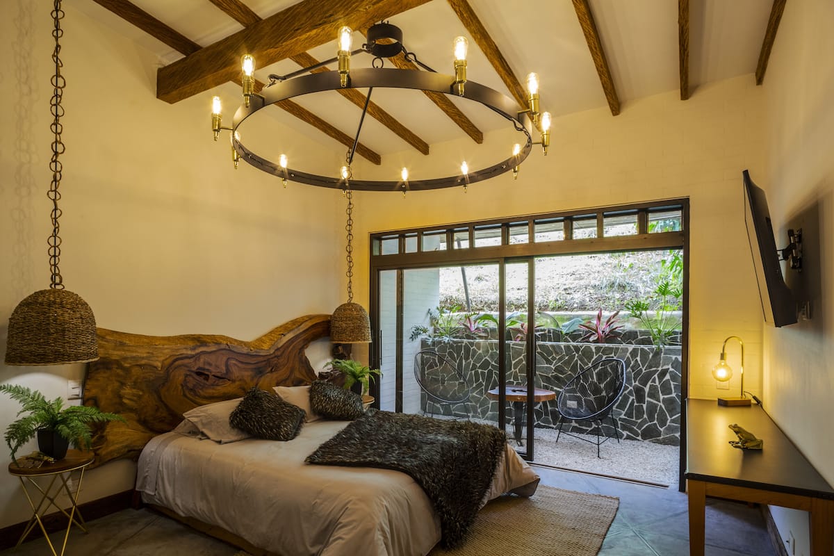 A bedroom with a wooden headboard, a hanging light fixture, a bed with a woven blanket, and a view of an outdoor garden through a glass door.