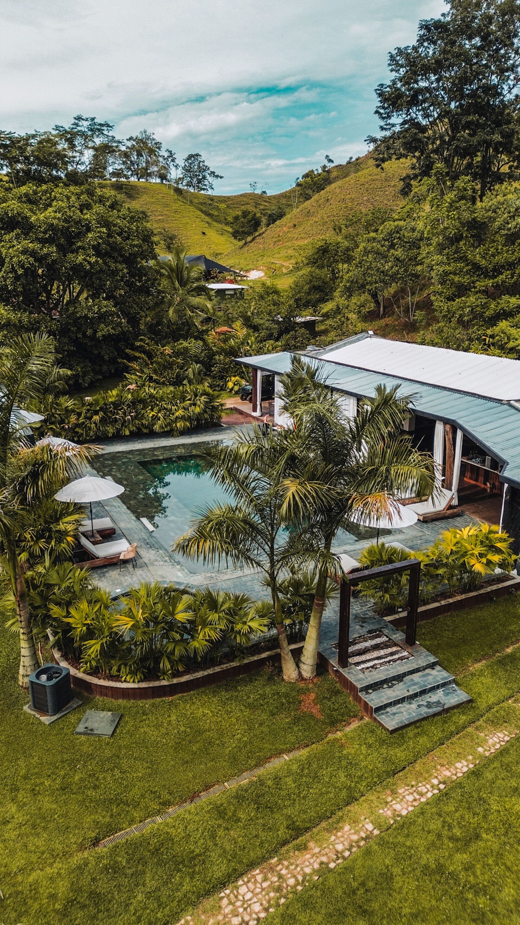 Una piscina grande rodeada de abundante vegetación, con una casa y un área de estar al aire libre al fondo.