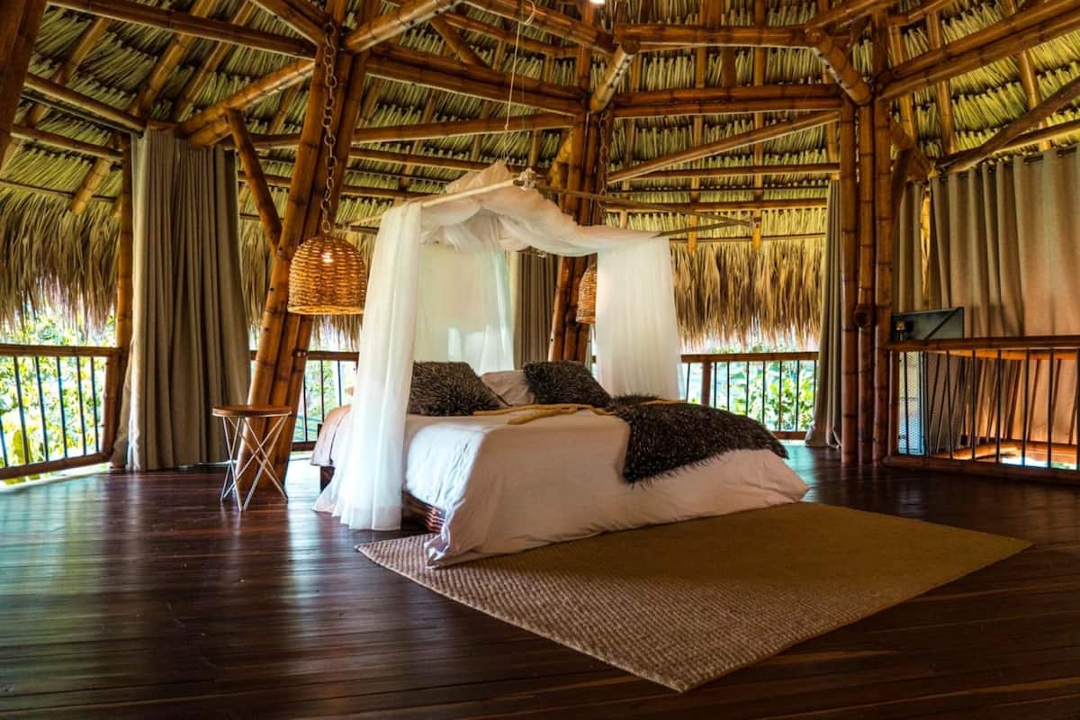 A room with a bed under a canopy, wooden flooring, and a view of greenery outside.