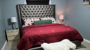 Ein Schlafzimmer mit einer gesteppten Kopfteile, einer roten Bettdecke, einem flauschigen weißen Pouf und einem Nachttisch mit Lampe.