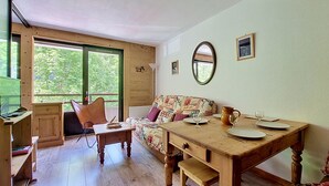 A cozy room with a wooden table, a sofa with patterned cushions, a mirror on the wall, and a window with a view of greenery.