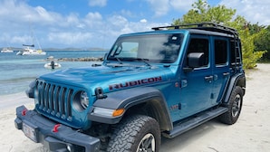 A blue Rubicon Jeep Wrangler parked on a sandy beach with a clear view of the ocean and boats in the background.