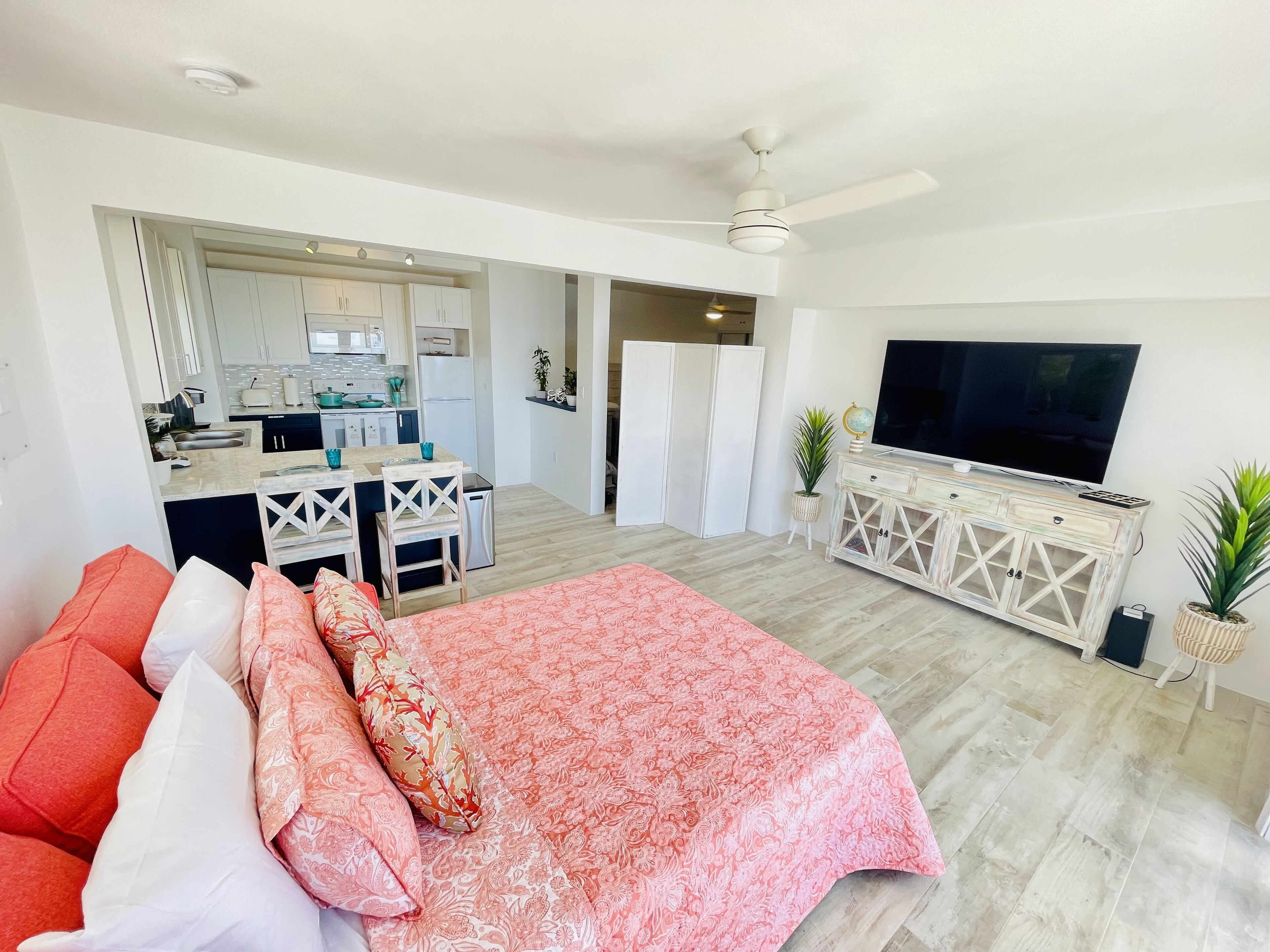 A modern living room with a large bed, a TV, and a kitchen in the background.
