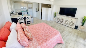 A modern living room with a large bed, a TV, and a kitchen in the background.