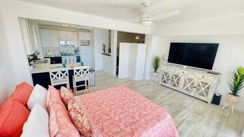 A modern living room with a large bed, a TV, and a kitchen in the background.