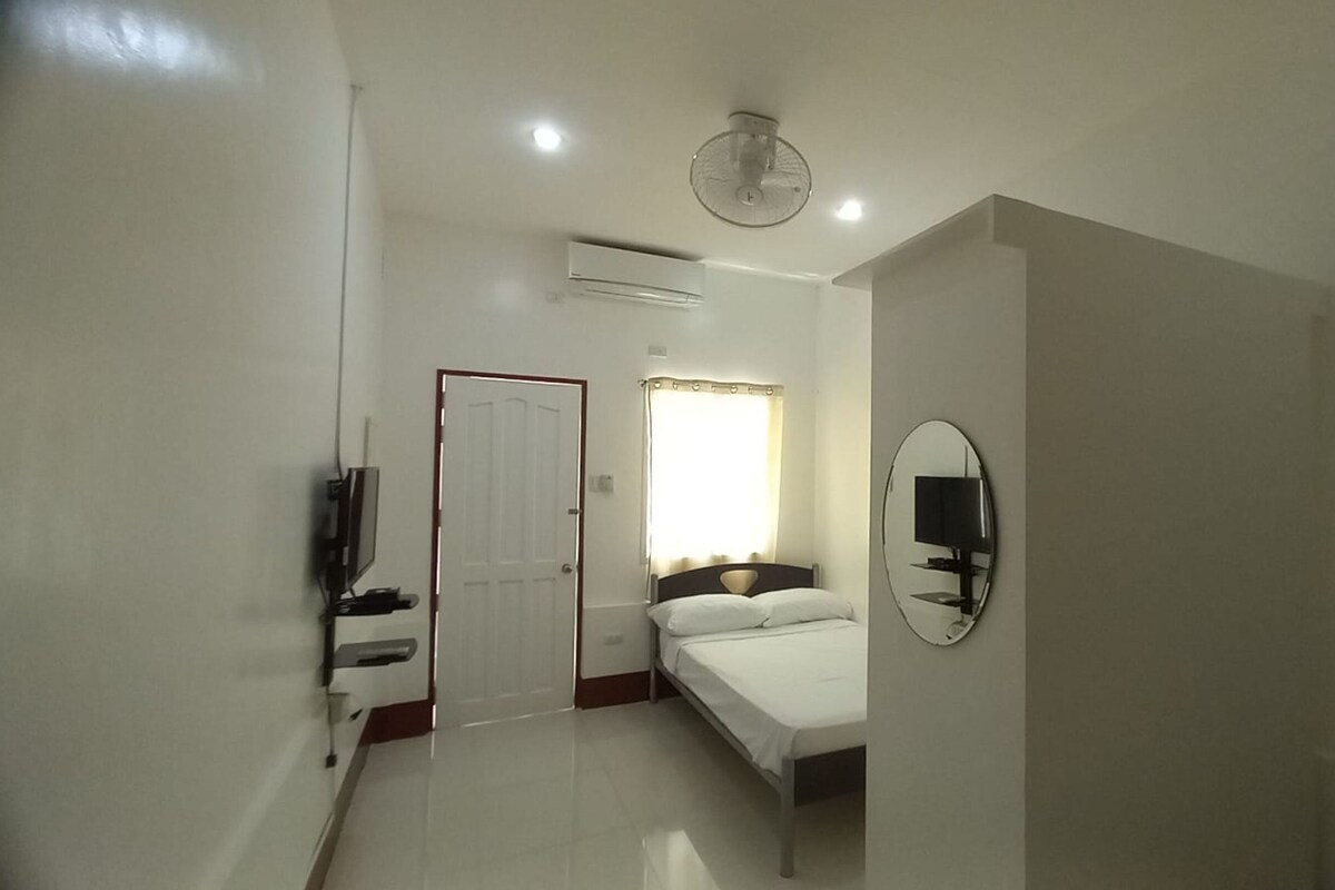 A bedroom with a bed, a television, an air conditioning unit, a door, and a window with curtains.