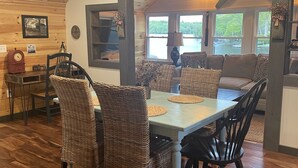 A dining area with a wooden table and wicker chairs, a ceiling fan, and a view of a lake through large windows. - Lakefront Bungalow (Otis)