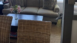 A cozy living room with a wooden ceiling, a beige sofa with patterned cushions, a vase of flowers, and a wicker coffee table. - Lakefront Bungalow (Otis)