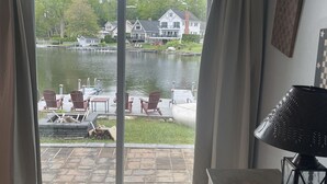 A room with a sliding glass door leading to a deck with outdoor furniture and a view of a lake with houses and docks. - Lakefront Bungalow (Otis)