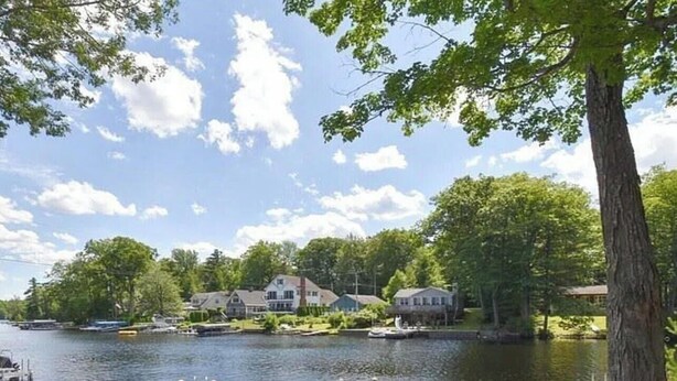 A lakeside deck with Adirondack chairs and a fire pit, overlooking a calm lake with boats and houses in the background. - Lakefront Bungalow (Otis)