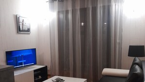 Un soggiorno con una TV a schermo piatto, un tavolino da caffè bianco, un divano in pelle nera e una finestra con tende trasparenti.