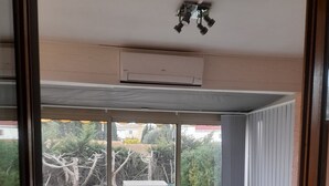 Una veranda con vetrate che presenta un tavolo rotondo e sedie, una panca integrata e vista su un giardino retrostante con una recinzione bianca e alberi.