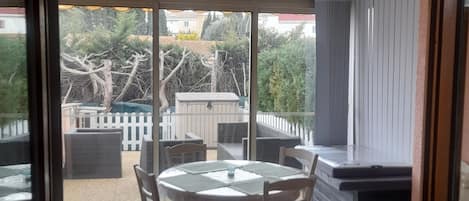 A glass-enclosed patio with a round table and chairs, a built-in bench, and a view of a backyard with a white fence and trees.