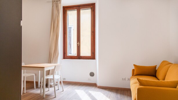 A room with a wooden floor, a white table with two chairs, a yellow sofa, a window with a wooden frame, and a white wall. - Cozy Studio in the heart of Navigli (Milano)