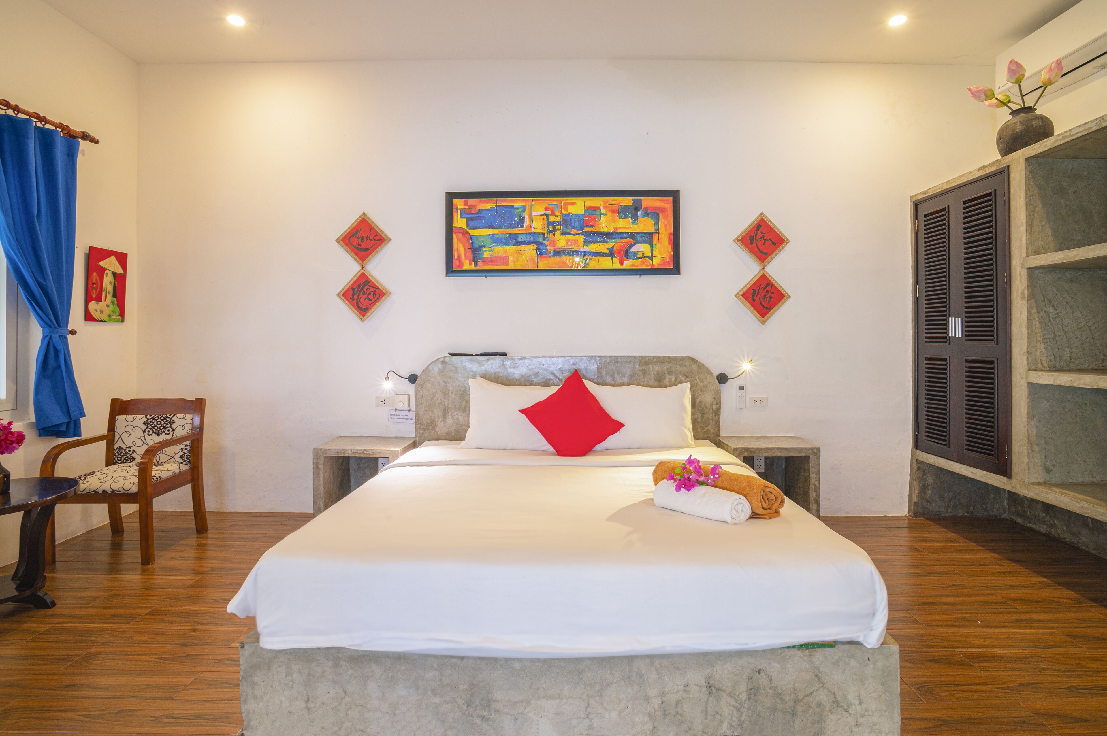 A hotel room with a large bed, a wooden chair, a small table with a vase of flowers, and a wardrobe.