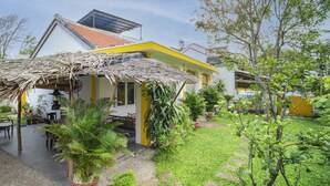 A house with a thatched roof, yellow exterior walls, and a garden with various plants and a small patio area.