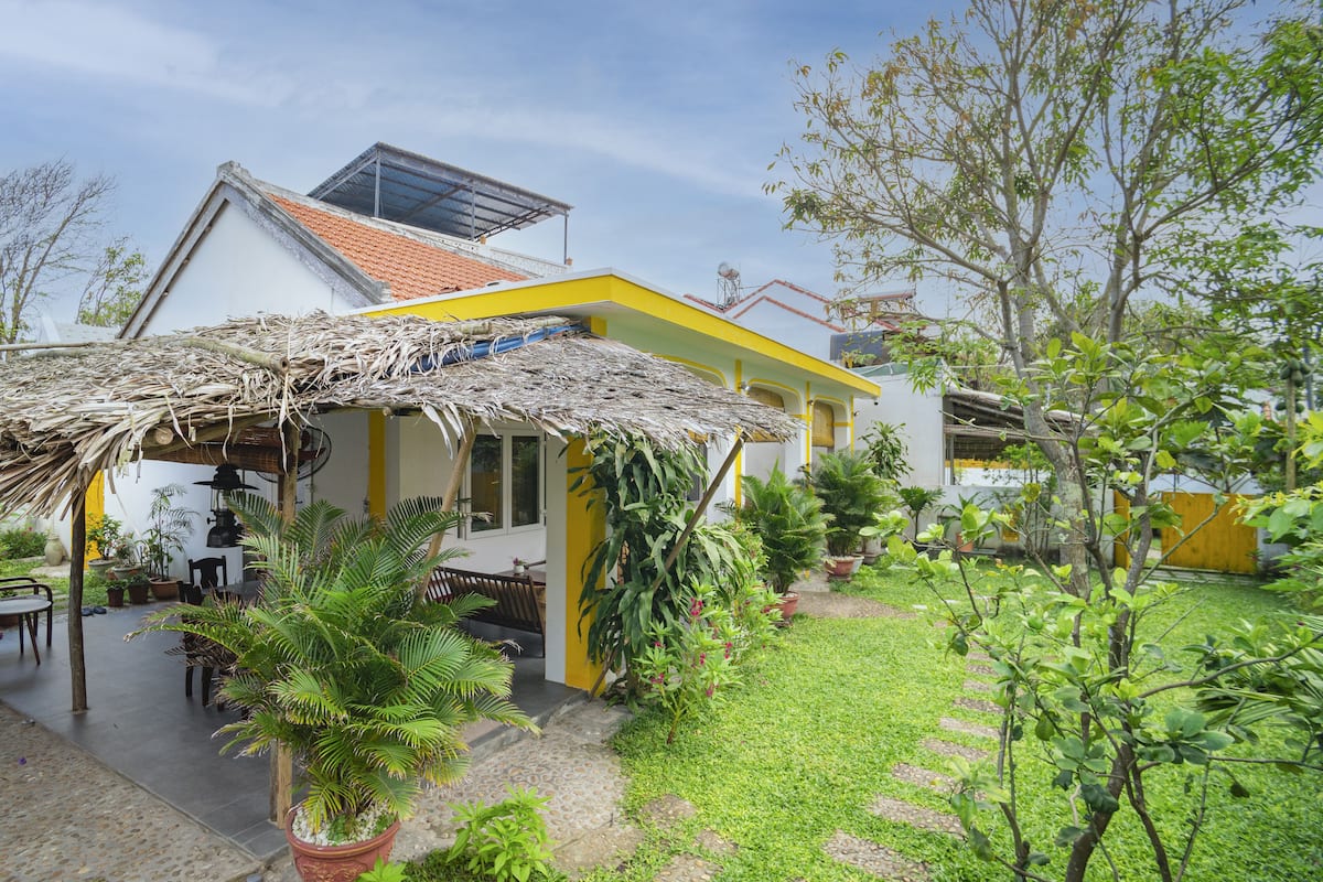 A house with a thatched roof, yellow exterior walls, and a garden with various plants and a small patio area.