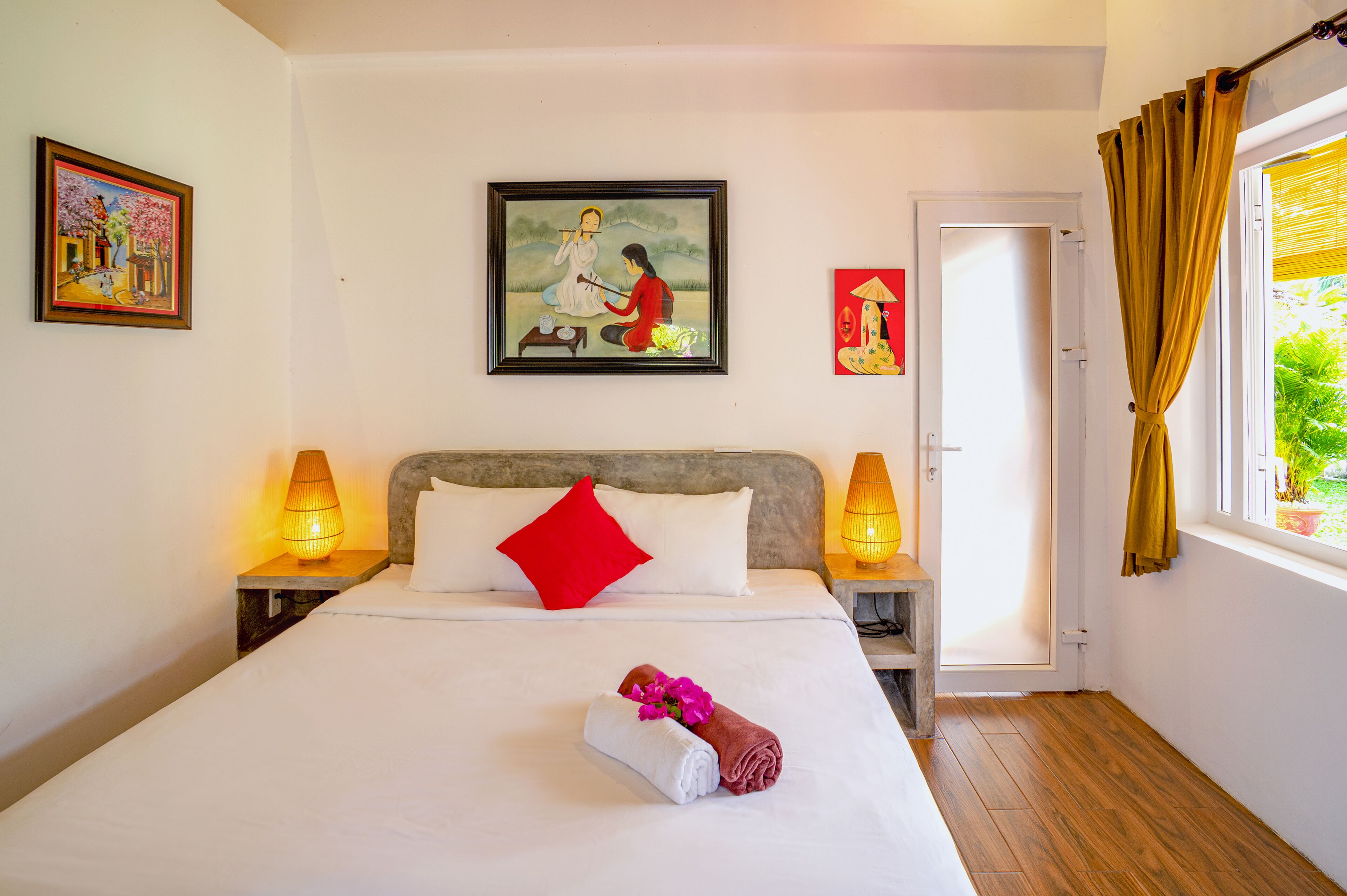 Una cama bien hecha con un almohadón rojo, dos lámparas de noche y un cuadro enmarcado encima.