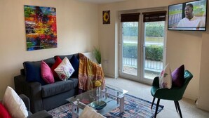 A living room with a dark sofa, colorful cushions, a glass coffee table, a green chair, a large abstract painting, a sunflower picture, and a TV on the wall. - Exquisite Modern Homely Flat (Cambridge)