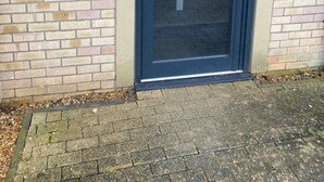 A building entrance with a glass door, a metal plaque indicating the property is managed by INCOR, and a security keypad on the wall. - Exquisite Modern Homely Flat (Cambridge)