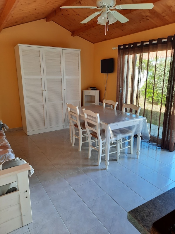 Un comedor con mesa y sillas, ventilador de techo, televisor y vistas a zonas verdes.