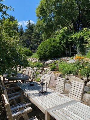 An outdoor seating area with wooden tables and chairs, surrounded by rocks and greenery.