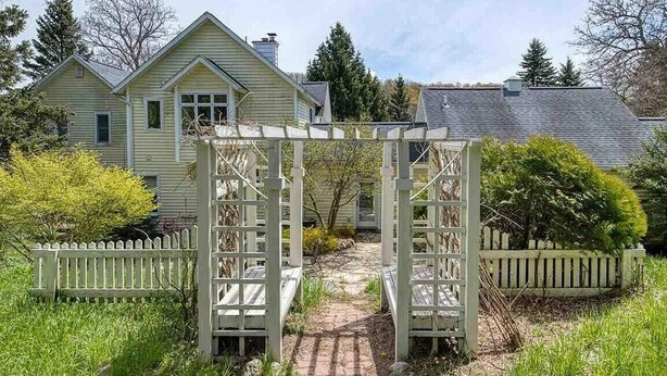 A wooden arbor with a brick pathway leading to a house with a gabled roof and a picket fence.