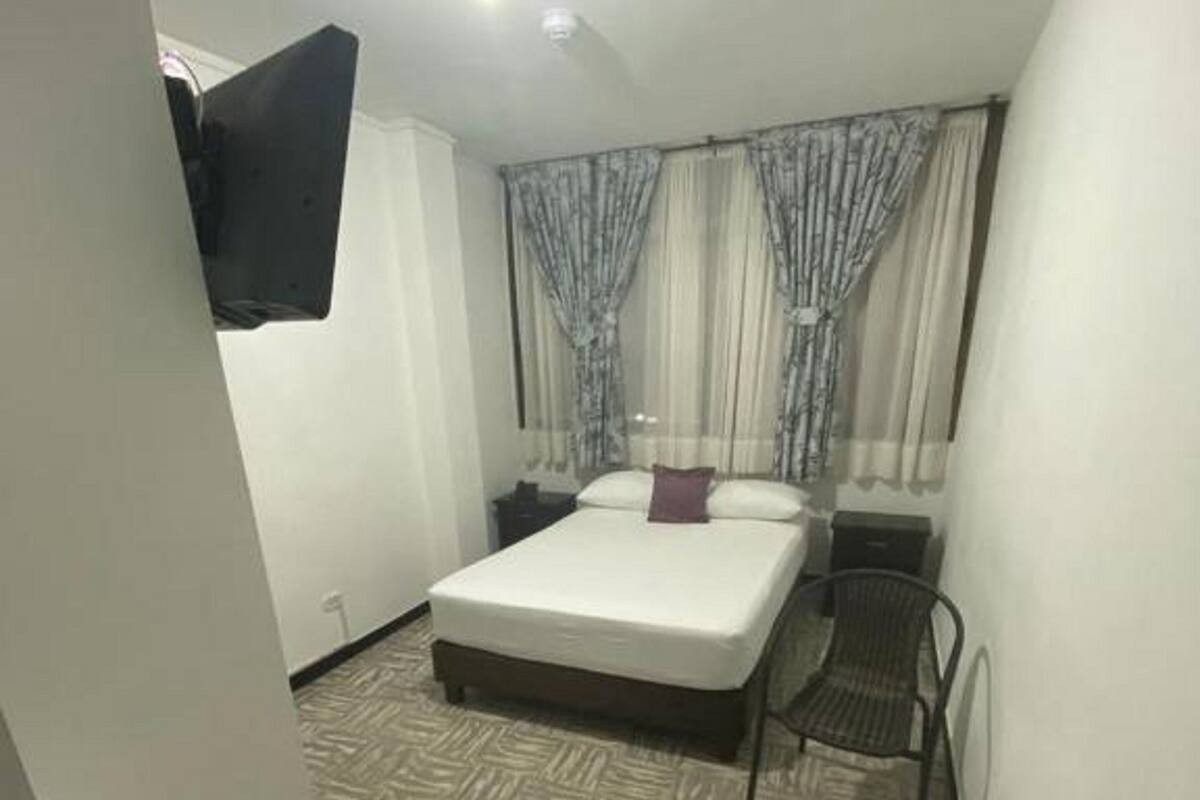A bedroom with a bed, a chair, a nightstand, curtains, and a mounted TV.