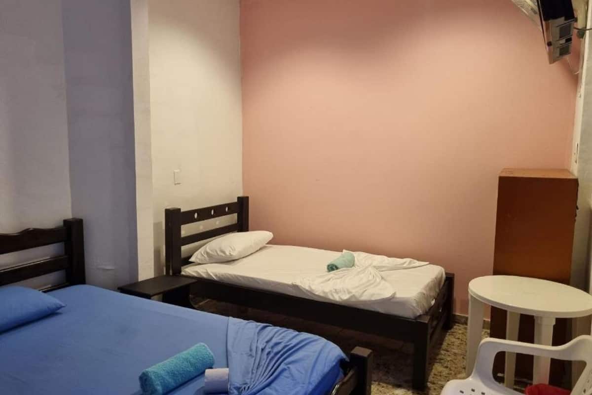 A room with two beds, one with blue bedding and the other with white bedding. There is a small white chair and a wooden cabinet.