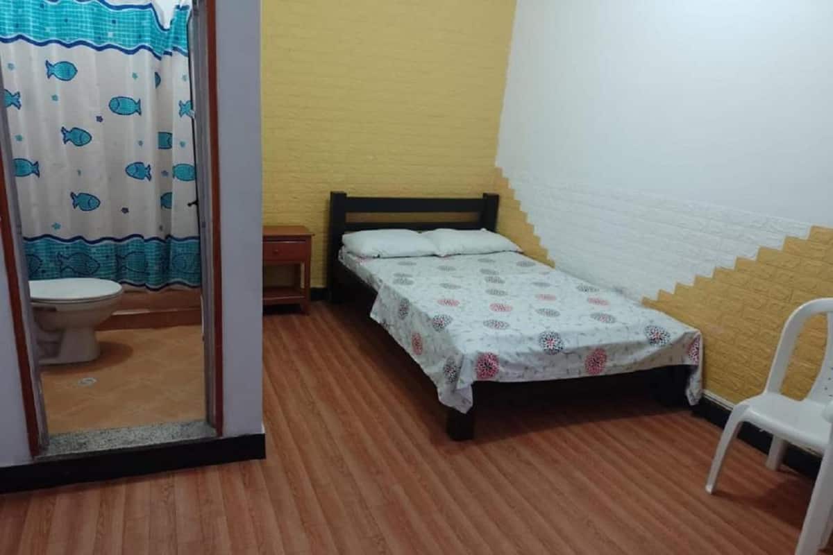 A small, single-room accommodation with a wooden floor, a bed with a patterned bedspread, a white chair, and a bathroom with a toilet and a shower curtain.
