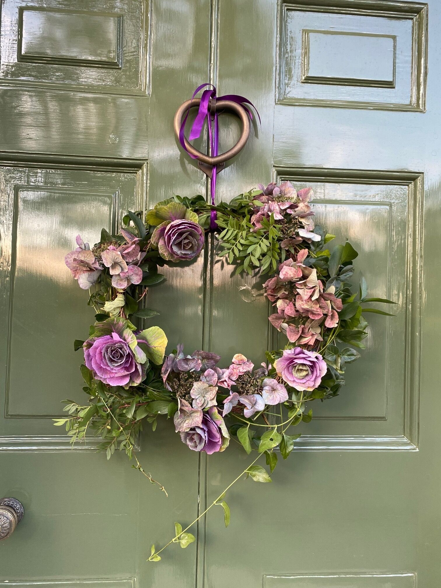 A green door with a heart-shaped wreath made of flowers and greenery.