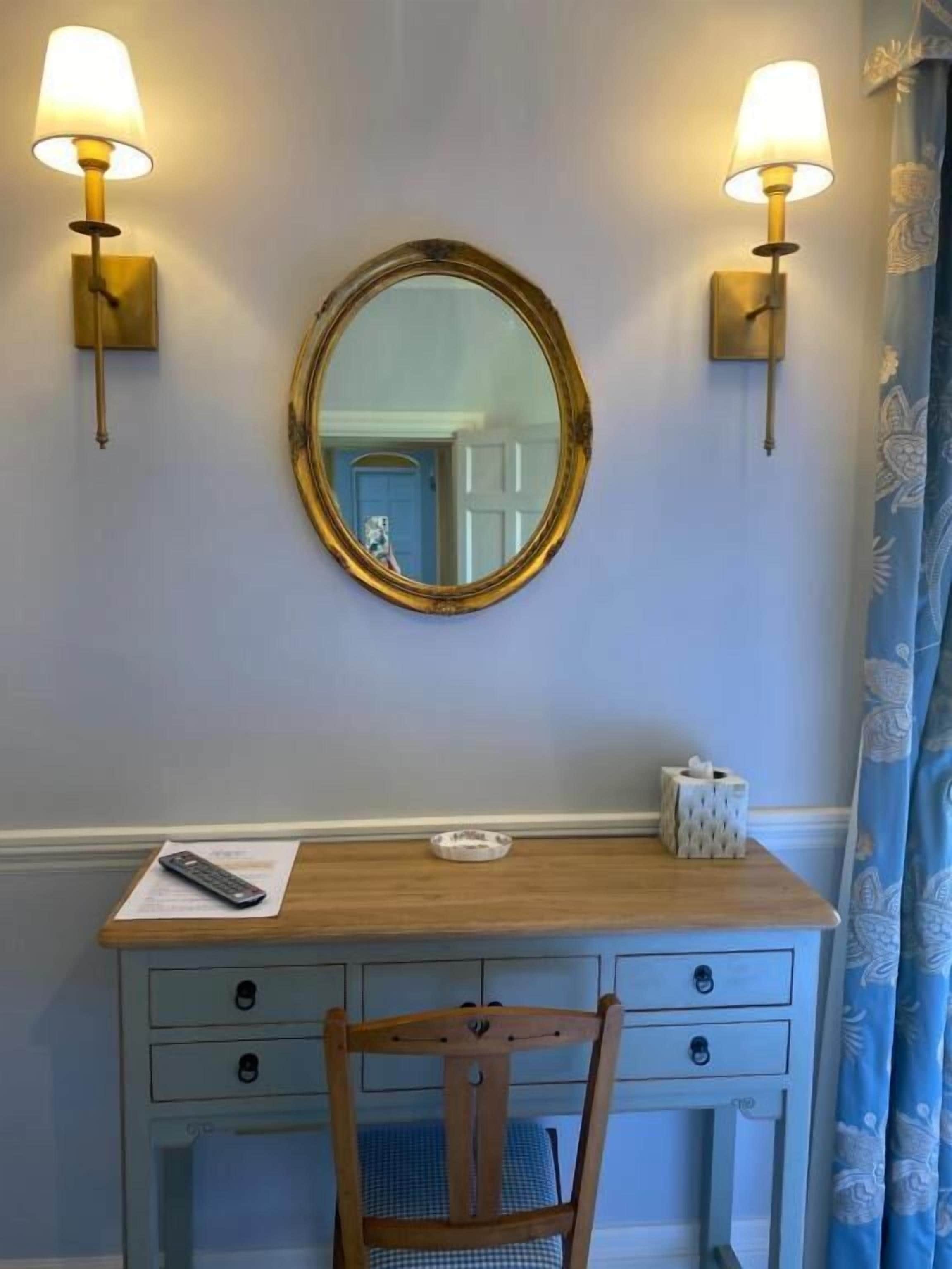 A wooden desk with a round mirror, two wall-mounted lamps, a tissue box, and a remote control on the desk.