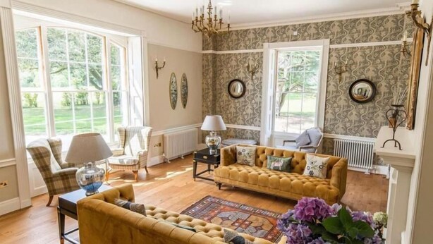A spacious living room with two yellow sofas, a coffee table, and a chandelier. There are large windows and a fireplace. - Reymerston Hall (Norwich)