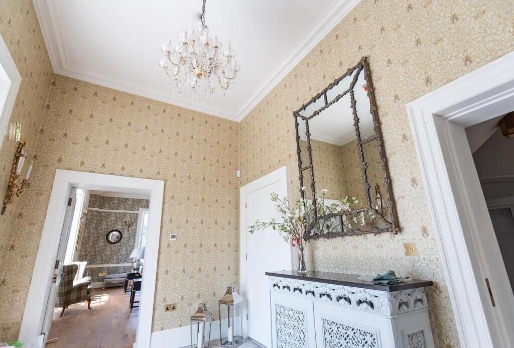 A room with patterned wallpaper, a chandelier, a mirror, and a cabinet with decorative items.