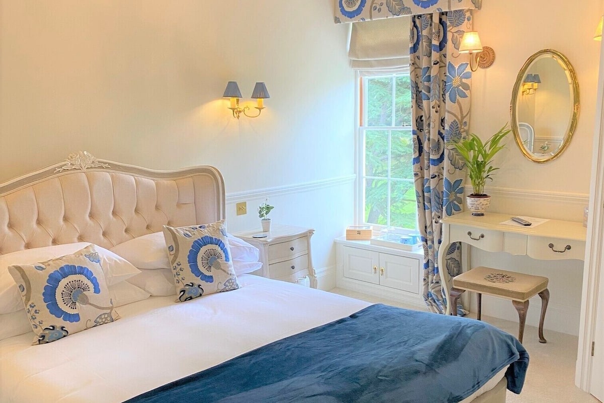 A bedroom with a large bed, a chandelier, a dresser, a mirror, and a window with curtains.