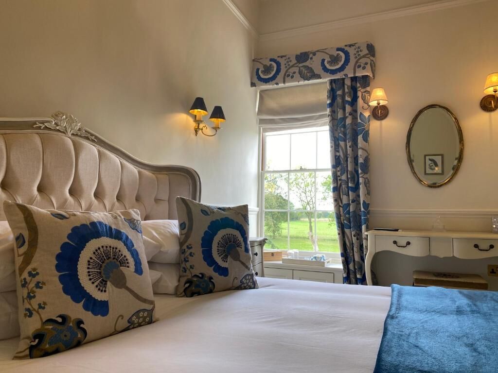 Un dormitorio con cabecera capitonada, dos almohadones decorativos, una mesita de noche, un aparador, un espejo y una ventana con cortinas.